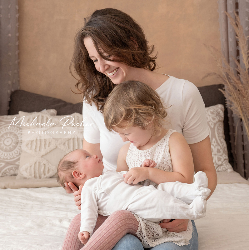 Mama sitzt mit ihrer kleinen Tochter auf dem Schoß, während diese das Baby liebevoll hält; beide Kinder tragen helle Kleidung und blicken sich sanft an.